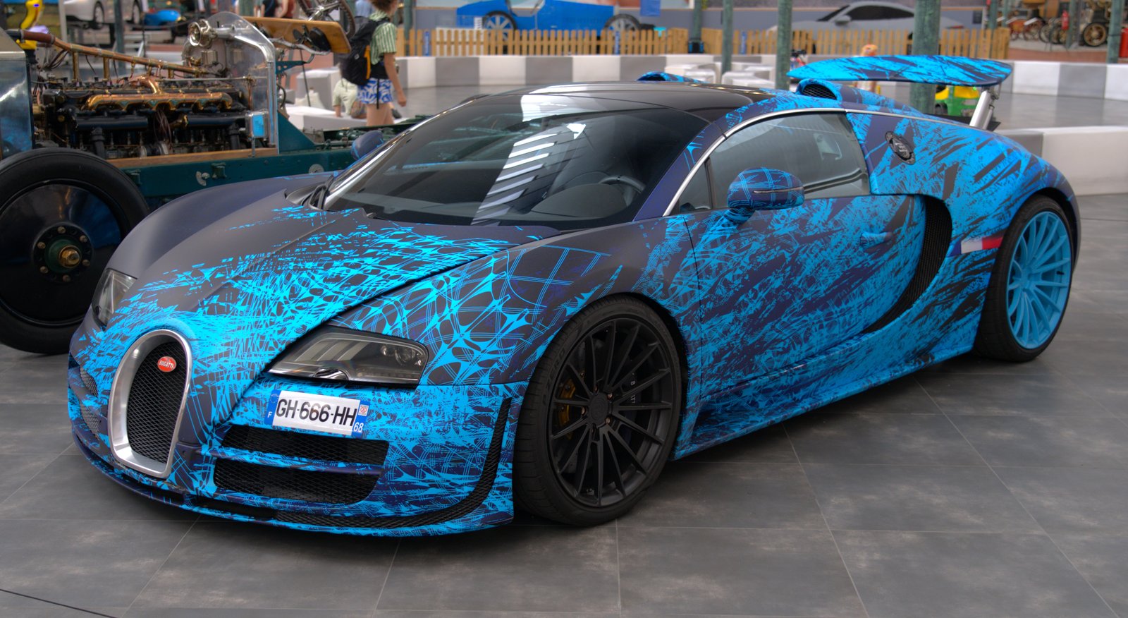 A Bugatti Veyron wrapped in electric-blue brushstroke livery stands on a grey tiled museum floor; behind it on the left an exposed early-1900s car chassis and engine, and a visitor walking past in the background.