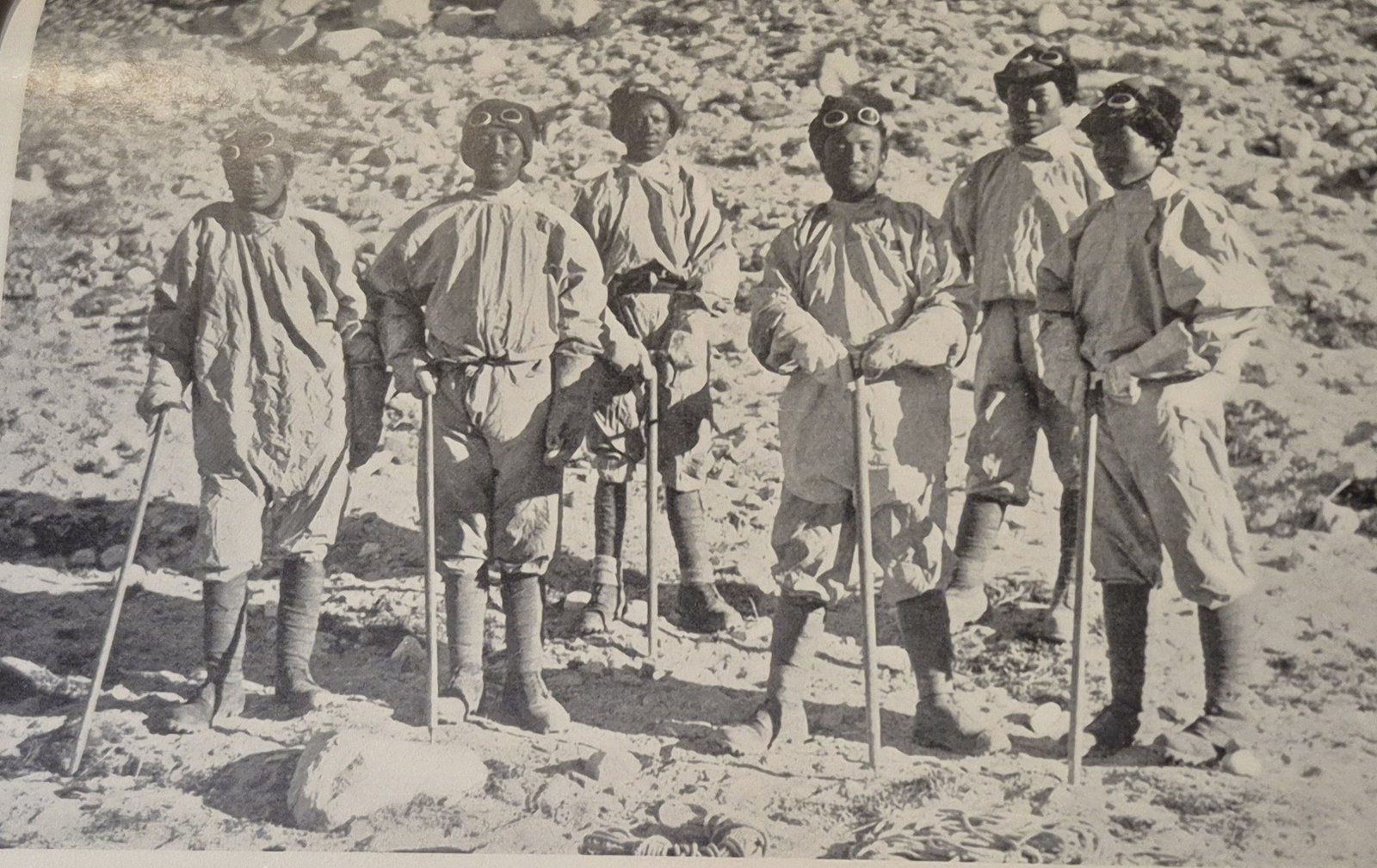An old black-and-white photograph: six Sherpa climbers in loose windproof suits, snow goggles pushed up on their caps, stand in a row on rocky ground holding ice axes.