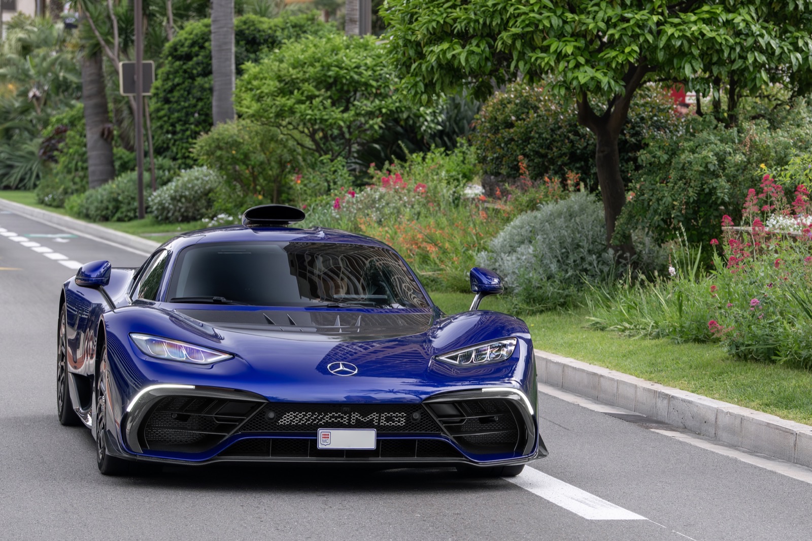 A blue Mercedes-AMG ONE hypercar, low and wide with a roof air scoop, driving toward the camera on an ordinary two-lane road lined with palms and flowering gardens.