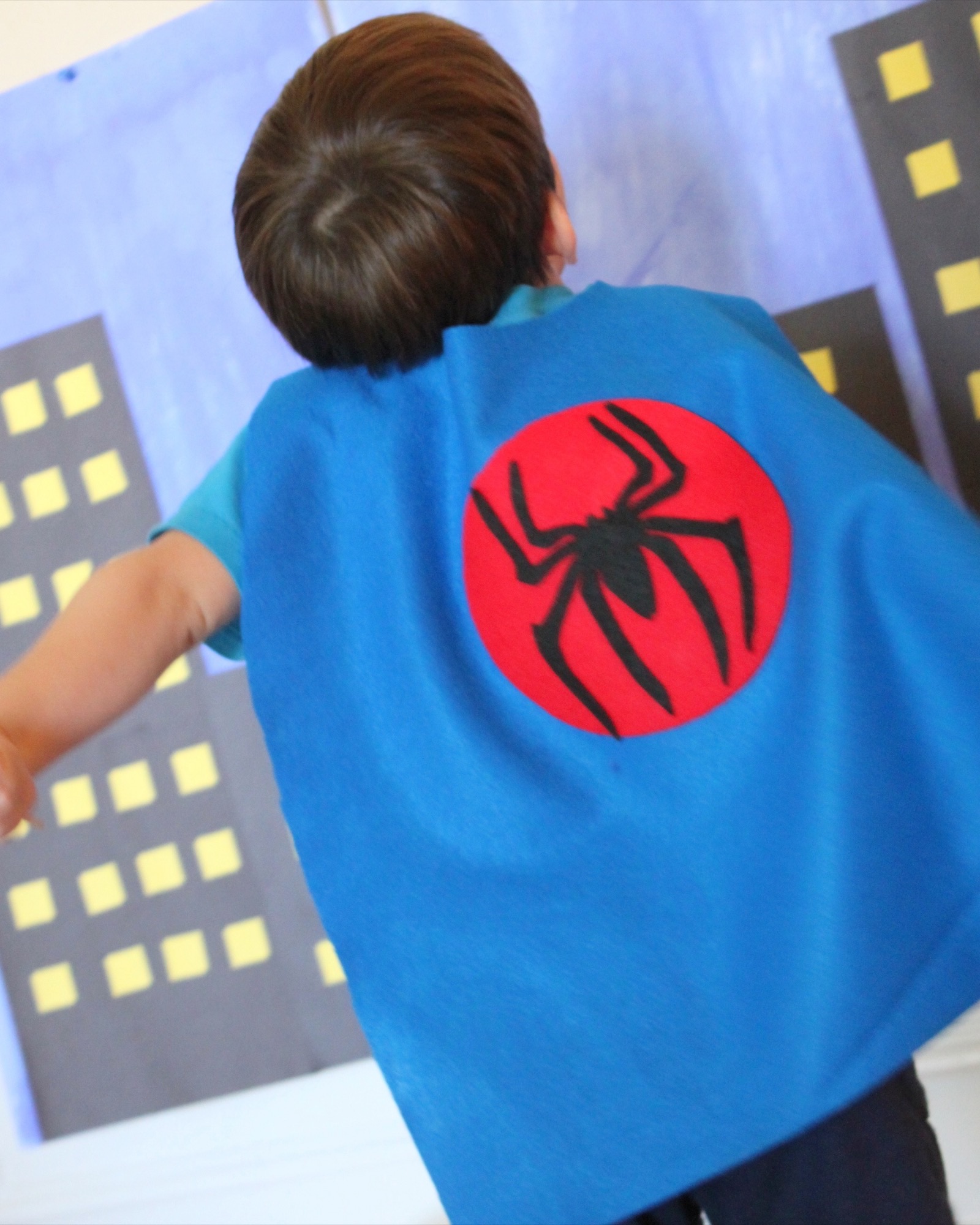 A small boy seen from behind in a blue felt cape with a red circle and black spider emblem, one arm out, facing a painted cartoon city skyline with yellow windows.