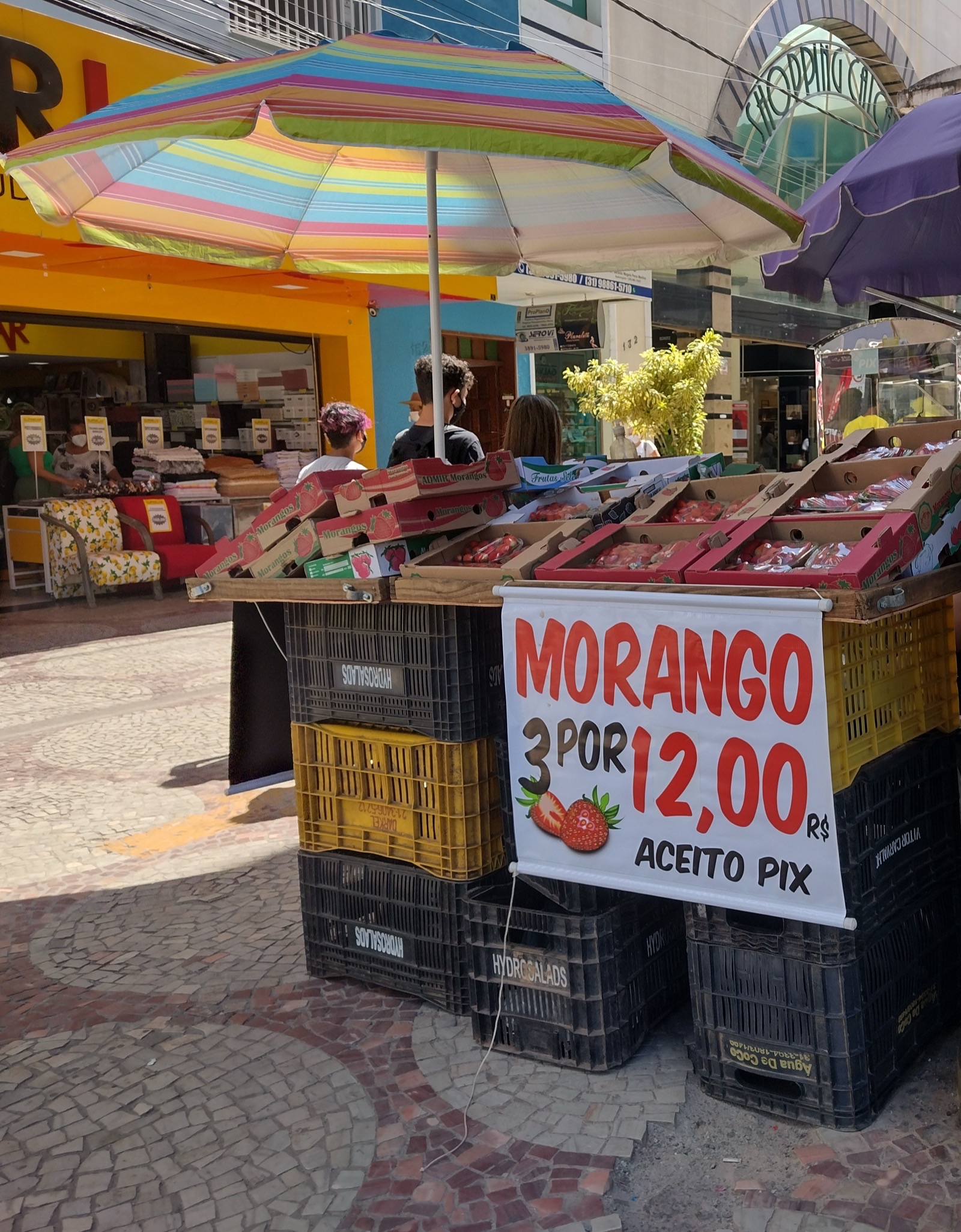 A street stall of strawberry boxes stacked on plastic crates under a striped umbrella in a Brazilian shopping street. A hand-lettered sign reads 'Morango 3 por 12,00 R$, aceito Pix'.