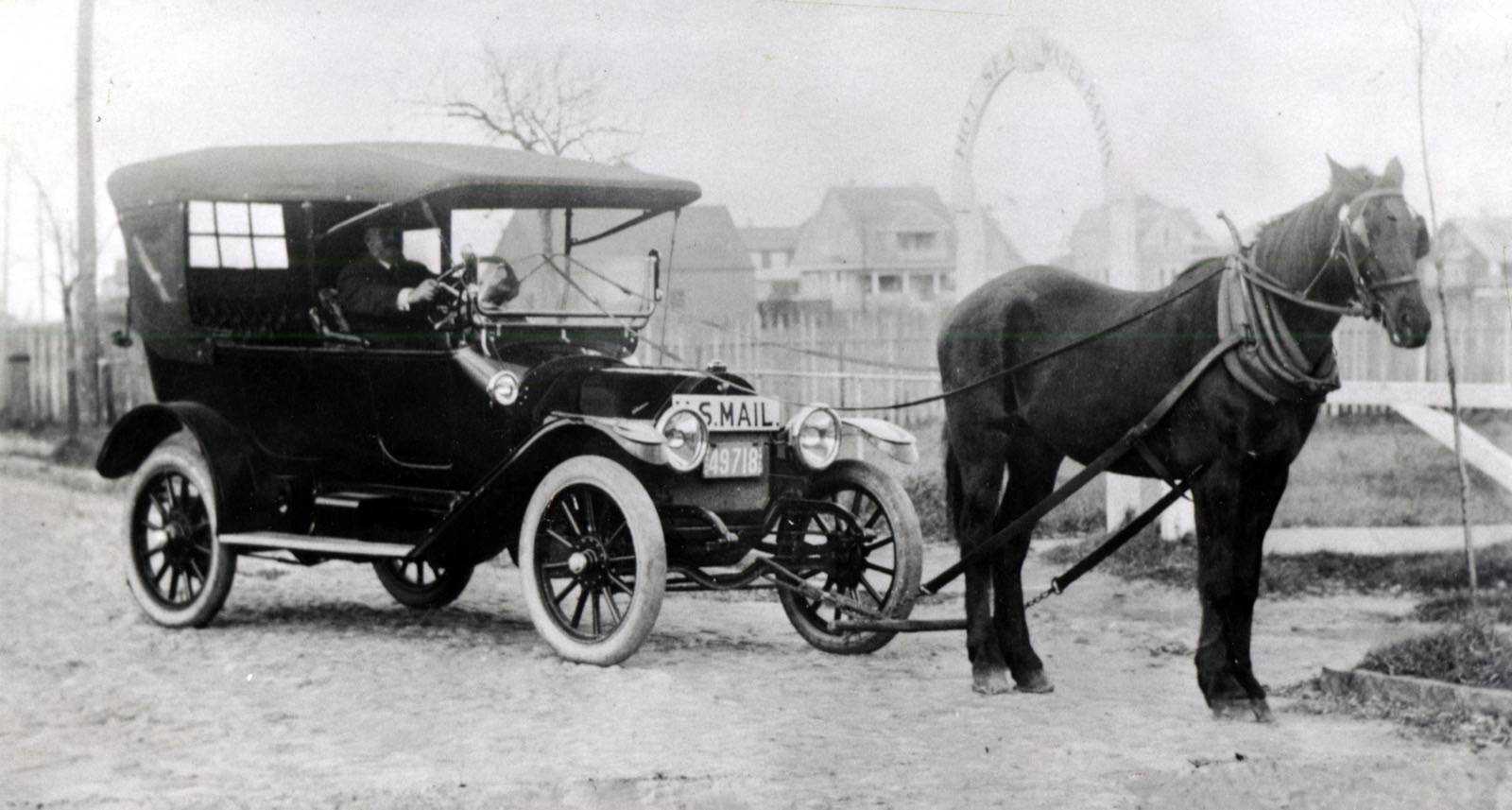 Black-and-white photo of an early touring car marked 'U.S. Mail', a man at the wheel, hitched by long poles to a dark horse that is towing it along an unpaved street.