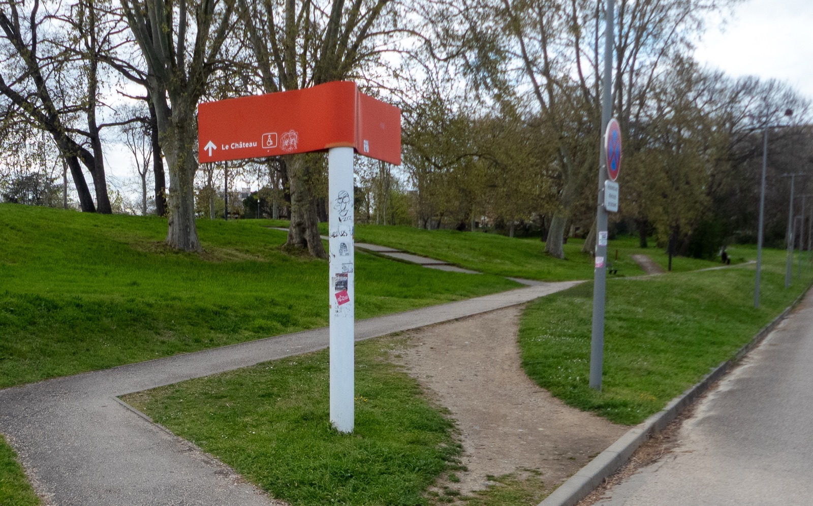 A paved footpath curves up a grassy slope past a red campus signpost; beside it, a bare dirt track worn by walkers cuts its own line across the grass.