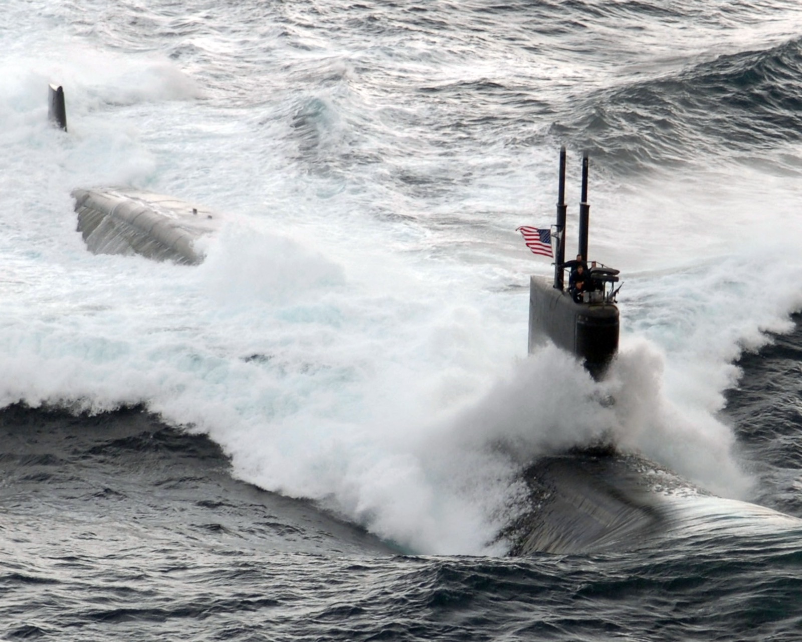 A US Navy attack submarine running fast on the surface, its hull buried in a wall of white spray; only the sail, with a US flag and sailors on the bridge, and the tail fin show above the wave.